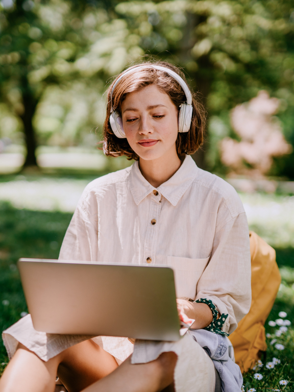 Frau sitzt mit Kopfhörern und mit Laptop in der Natur